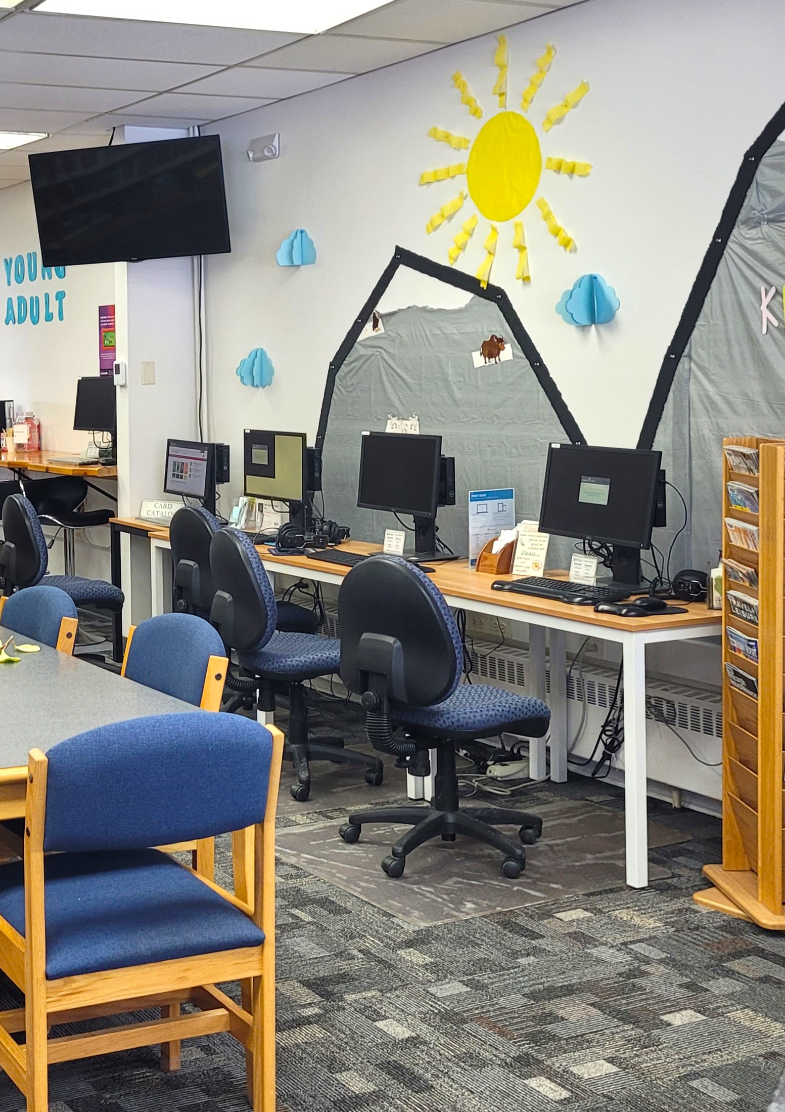 A library area with computer stations and blue chairs, featuring wall decorations of a paper sun, clouds, and mountain.