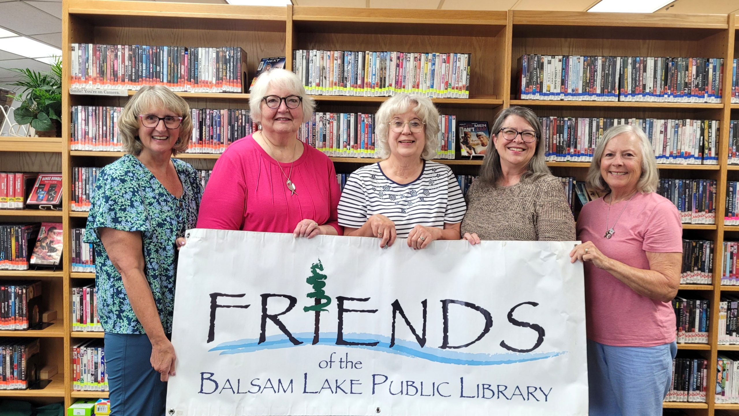 Five women stand together holding a banner that says "FRIENDS of the Balsam Lake Public Library," with a bookshelf of DVDs in the background.